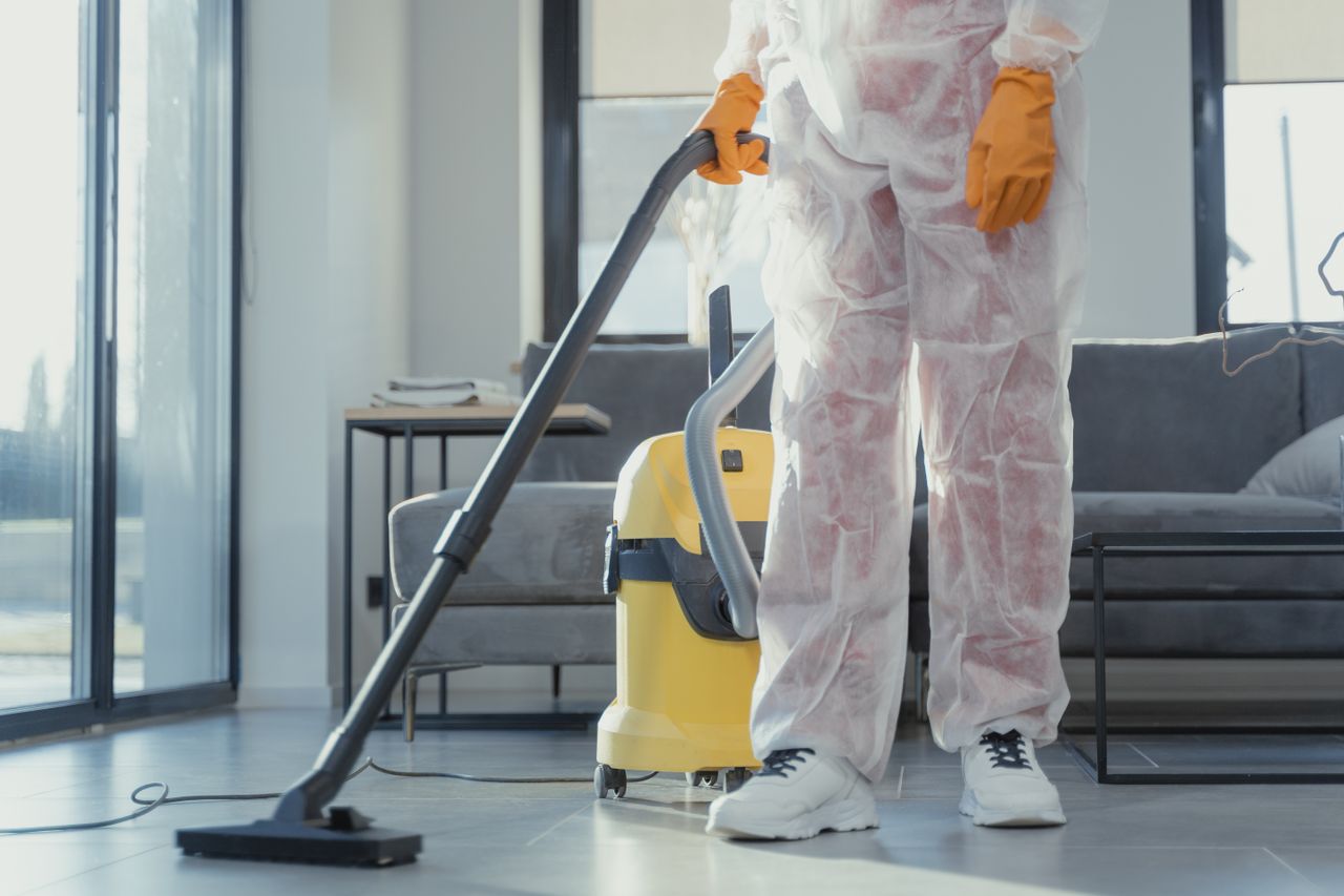 Cleaner v ochranném overalovém oděvu a rukavicích vysává moderní obývací pokoj.